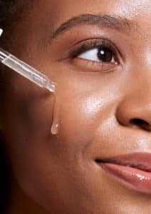 Black Woman, Dropper And Facial Skincare Cosmetics For Beauty, Hydration Or Oil Treatment For Healthy Face. Closeup Of African American Female Applying Serum To Cheek For Skin Hydrate, Glow Or Shine